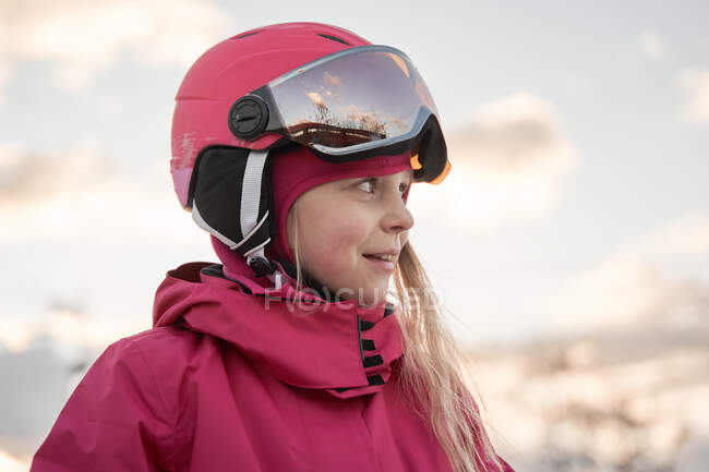 Веселая очаровательная девушка в розовой теплой спортивной одежде и шлеме стоит на просторной снежной местности и смотрит в сторону — стоковое фото