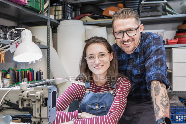Cheerful tattooed craftsman and charming seamstress looking at camera in messy workshop for creating motorcycle seat upholstery — Stock Photo