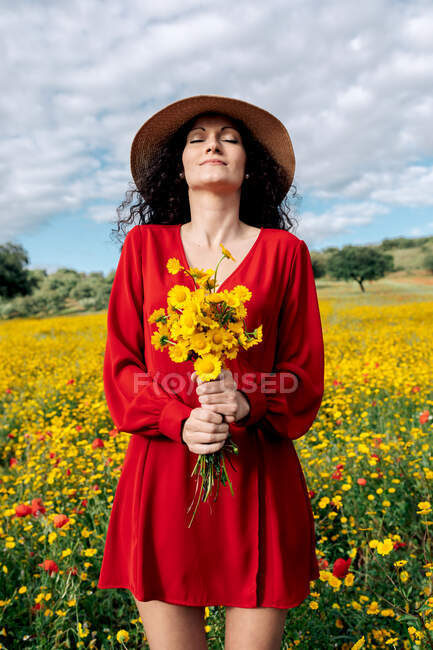 Femmina in cappello con gli occhi chiusi azienda fioritura fiori gialli nel campo di campagna sotto cielo nuvoloso — Foto stock