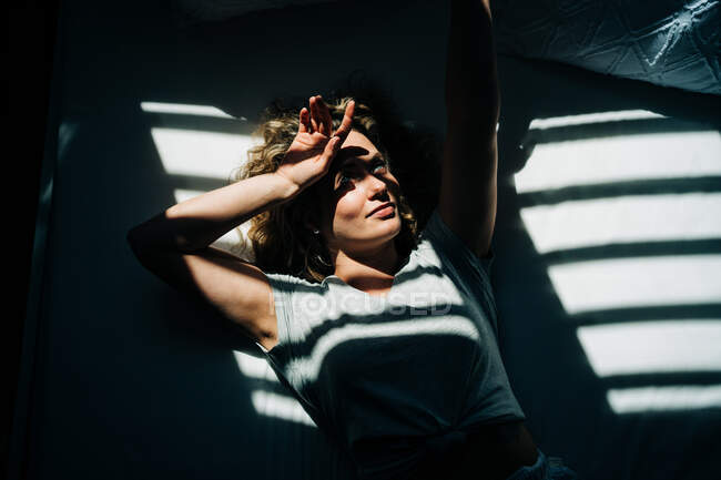 Зверху молода жінка з кучерявим світлим волоссям торкається чола з закритими очима, лежачи на зручному ліжку в сонячний ранок — стокове фото