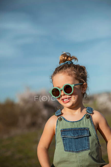 Маленькая девочка в комбинезоне и солнцезащитных очках, стоящая на лугу и наслаждающаяся летом в солнечный день в сельской местности — стоковое фото
