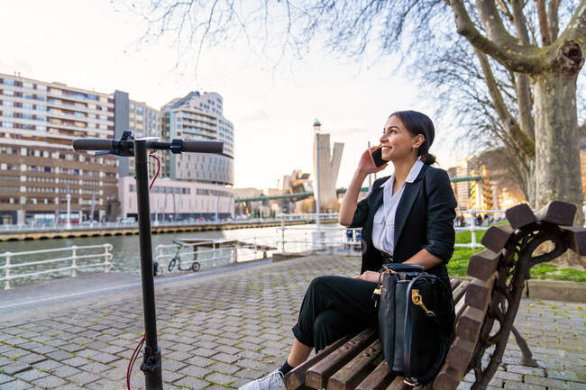 Sorrindo empresária étnica sentada no banco de madeira com pernas cruzadas falando no celular enquanto olha para o banco da cidade contra a scooter — Fotografia de Stock