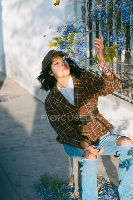 Young feminine ethnic female in stylish outfit sitting under blossoming tree branch on sunny day - foto de stock