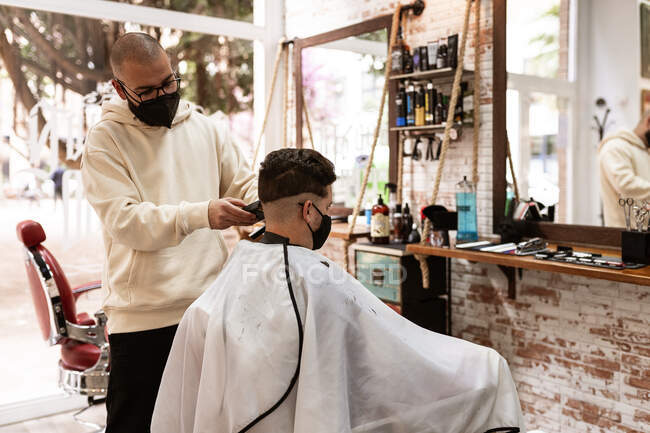 Männlicher Friseur mit Brille schneidet erwachsenen Kunden im Friseursalon während der COVID-19-Pandemie die Haare — Stockfoto
