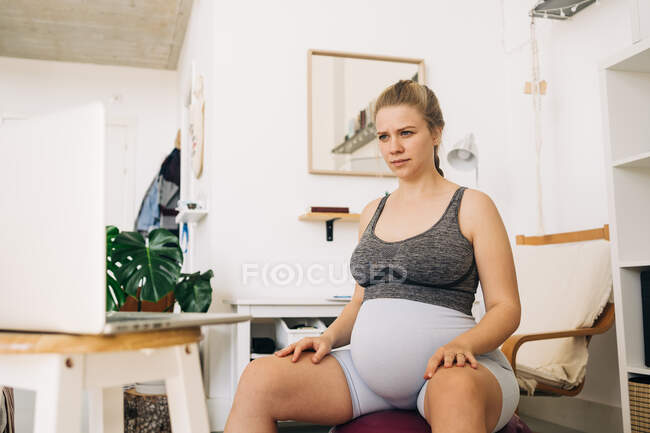 Joven mujer embarazada sentada en la pelota de ejercicio y portátil de navegación en la sala de estar - foto de stock