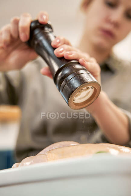 Low angle of crop female with grinder sprinkling uncooked poultry with black pepper while cooking at home — Stock Photo