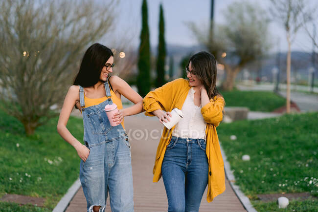 Content best female friends in casual wear with hot drinks in tumblers touching elbows while looking at each other in town — Stock Photo