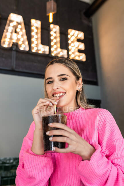 Веселая молодая женщина в розовом свитере, потягивающая холодный газированный безалкогольный напиток через солому, проводя свободное время в кафетерии и счастливо глядя в камеру — стоковое фото