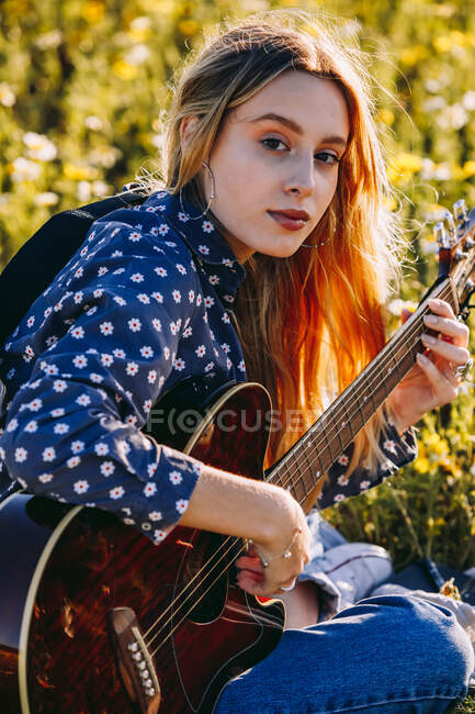 Jeune femme hipster réfléchie assise sur une prairie à la campagne écrivant des chansons sur un carnet et jouant de la guitare pendant la lumière du soleil d'été — Photo de stock