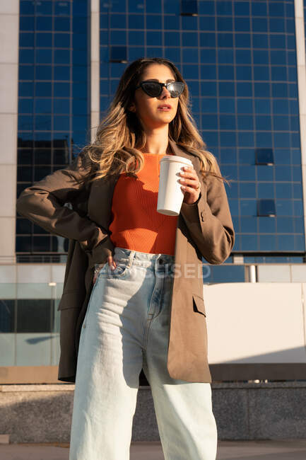 Молодая женщина в стильной одежде с горячим напитком, чтобы пойти и солнцезащитные очки с нетерпением жду в городе — стоковое фото