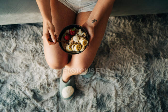 Top view anonymous female in shorts enjoying tasty oatmeal with ripe strawberries and sliced banana while sitting on bed — Stock Photo