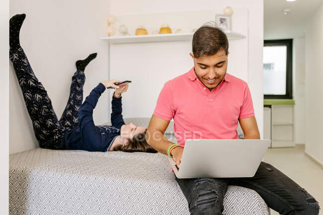 Щасливий молодий етнічний фрілансер, який працює дистанційно на ноутбуці, сидить на ліжку біля лежачої подруги, переглядаючи смартфон — стокове фото