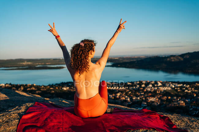Back view of happy topless female traveler with curly hair in leggings smiling and showing two fingers gesture with raised arms sitting on rocky cliff above coastal town — Stock Photo