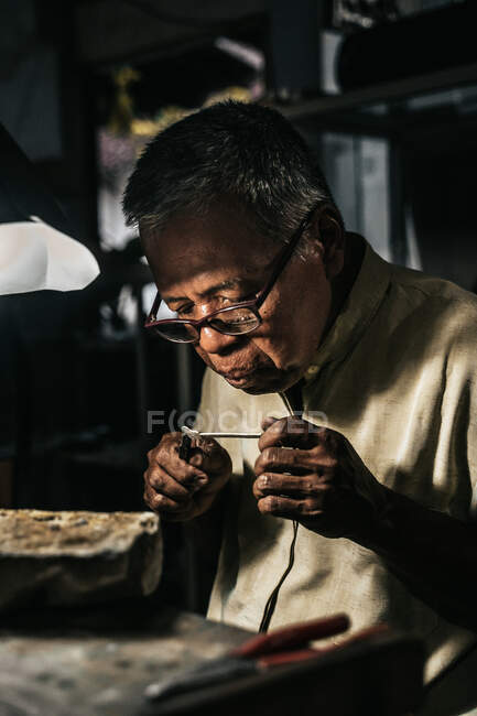 Concentrado senior afroamericano orfebre en ropa casual y gafas que sostiene la soldadura con alicates mientras crea joyas en el taller - foto de stock