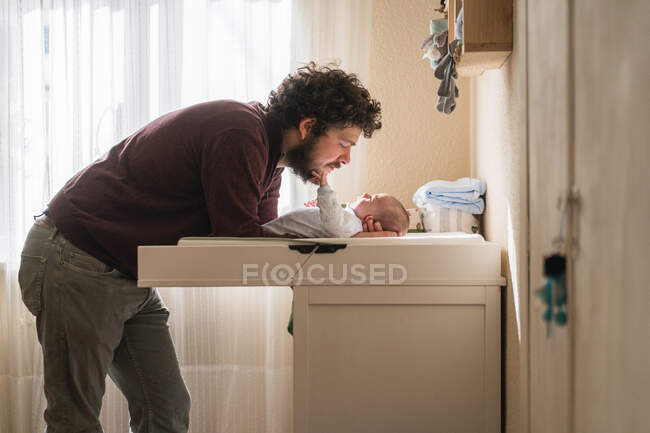 Side view of adult bearded dad with little child leaning forward at infant table while looking at each other indoors — Stock Photo