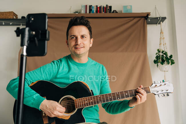 Веселый взрослый видеоблогер с гитарой и плектором, записывающий видео на мобильный телефон дома — стоковое фото