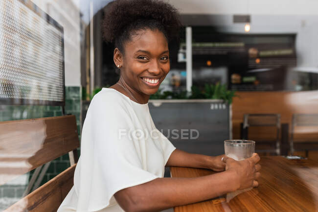 Seitenansicht einer ruhigen Afroamerikanerin in lässigem Outfit, die kaltes Süßwasser aus der Flasche in ein Glas mit Eis und Zitrone trinkt, während sie am hohen Tisch in der Cafeteria sitzt und in die Kamera schaut — Stockfoto