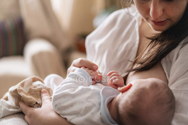 Cortada mãe irreconhecível em óculos amamentando anônimo criança sentada na sala da casa à luz do dia — Fotografia de Stock