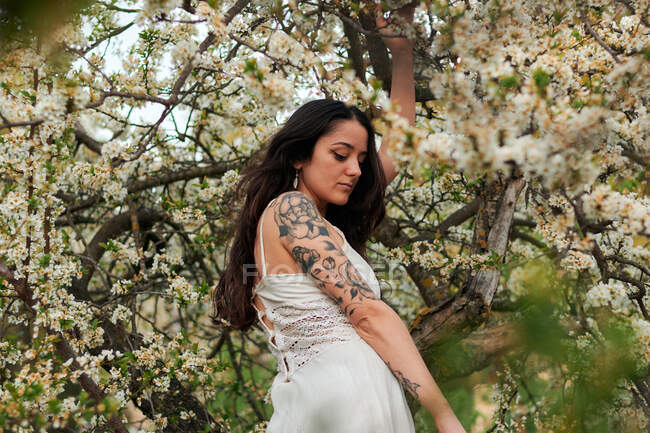 Young female with tattooed arm wearing white dress and standing in flowers of tree looking down — Foto stock