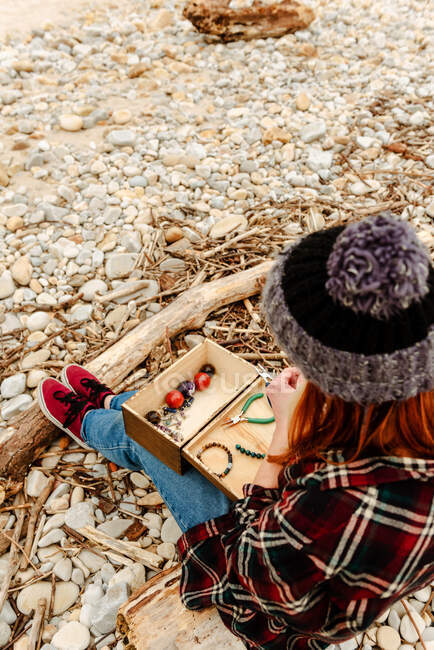 From above crop anonymous craftswoman creating handmade bijouterie while using semiprecious stones and sitting on beach near sea — Stock Photo