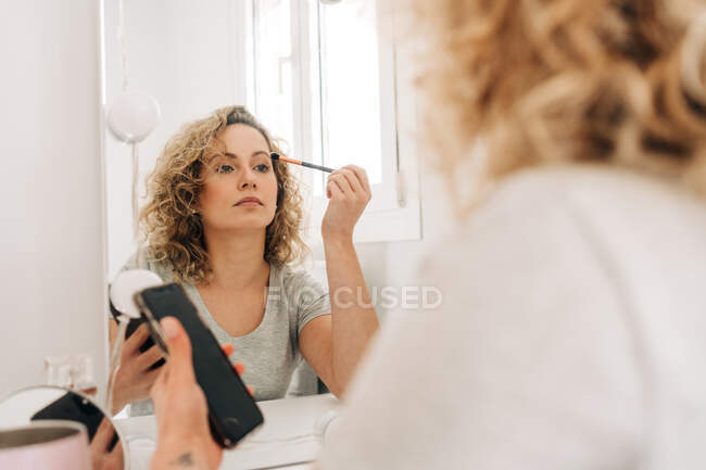 Mujer joven enfocada en camisa casual aplicando maquillaje mientras está sentada en la mesa de tocador con teléfono inteligente en el dormitorio ligero - foto de stock
