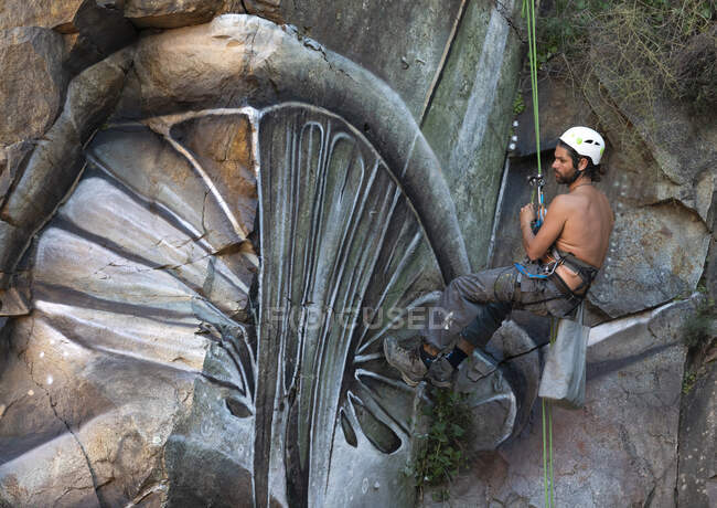 Vue de côté corps complet du peintre avec peinture en aérosol faisant graffiti accroché à la corde sur pente rocheuse raide — Photo de stock