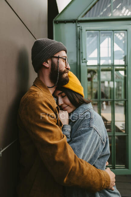 Вид збоку молода стильна пара м'яко обіймається, стоячи на дерев'яній терасі біля будинку — стокове фото