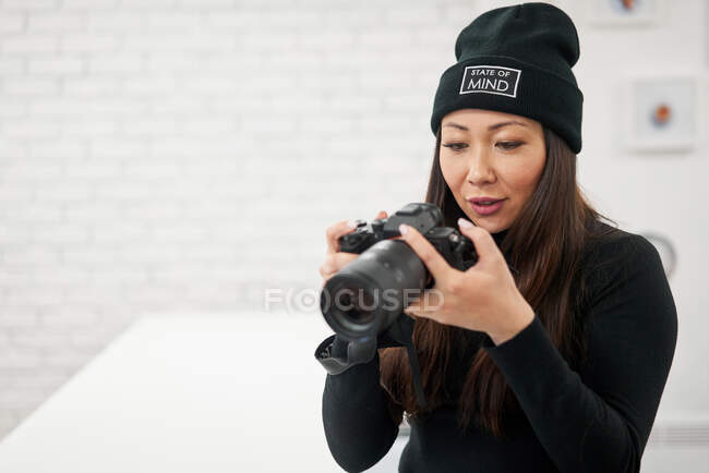 Donna etnica in berretto nero e camicia con fotocamera digitale su sfondo sfocato — Foto stock