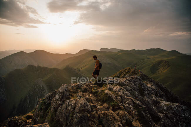 Vista laterale del giovane con zaino che cammina sulla cima rocciosa della montagna in valle contro il cielo del tramonto — Foto stock