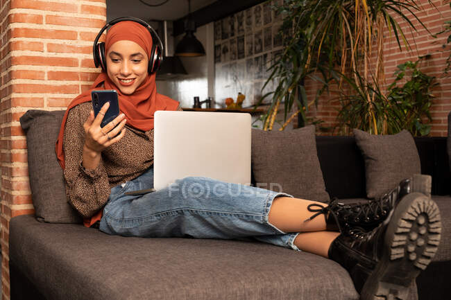 Longitud completa feliz joven musulmana femenina en ropa modesta hijab y auriculares inalámbricos sentados en acogedor sofá con netbook en vueltas y el teléfono inteligente de navegación - foto de stock