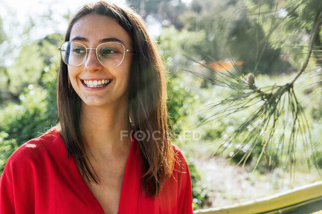 Jovem conteúdo feminino em óculos e roupas vermelhas olhando para longe no dia ensolarado no fundo borrado — Fotografia de Stock