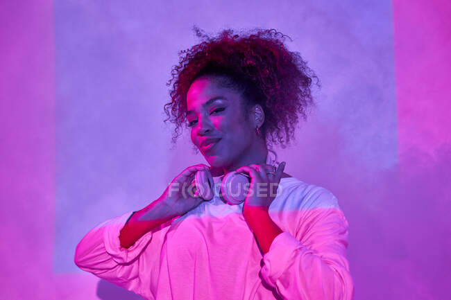 Porträt einer fröhlichen jungen Afroamerikanerin in weißen Kleidern, die Kopfhörer am Hals trägt und in die Kamera schaut, während sie im dunklen Studio im Neonlicht steht — Stockfoto