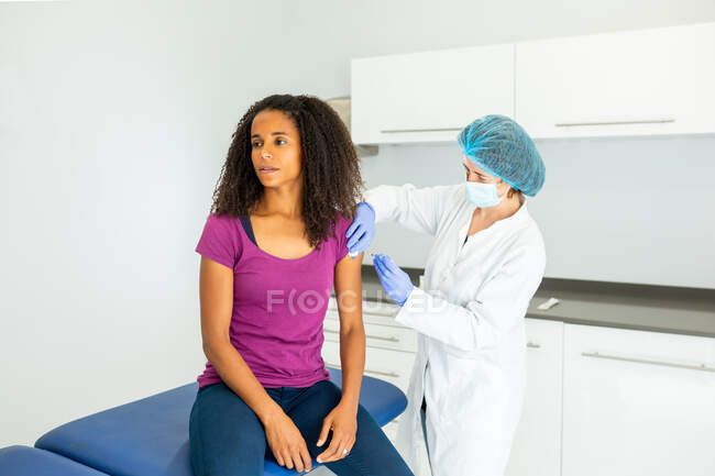 Female medical specialist in protective uniform, latex gloves and face mask vaccinating African American female patient in clinic during coronavirus outbreak — Stock Photo