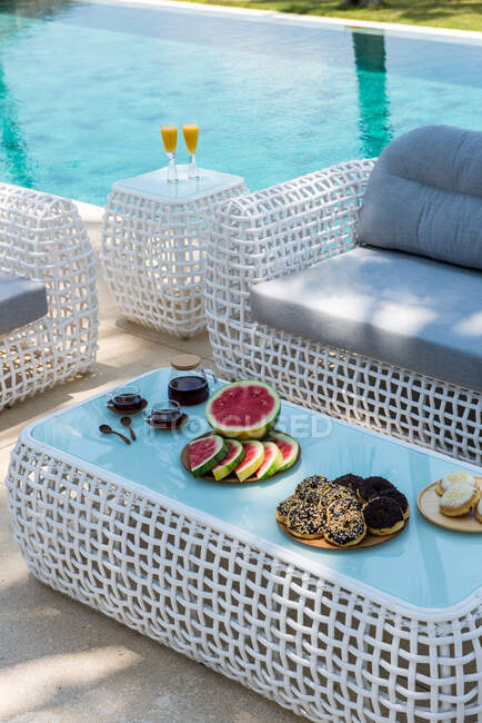 From above of plates with assorted appetizing pastries and fresh sliced watermelon served on table with cups of coffee near wicker sofa at poolside in tropical resort — Photo de stock