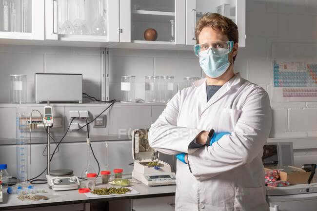 Химик-мужчина в стерильной маске, перчатках и защитных очках смотрит в камеру на анализатор влаги и аналитический баланс в лаборатории конопли — стоковое фото