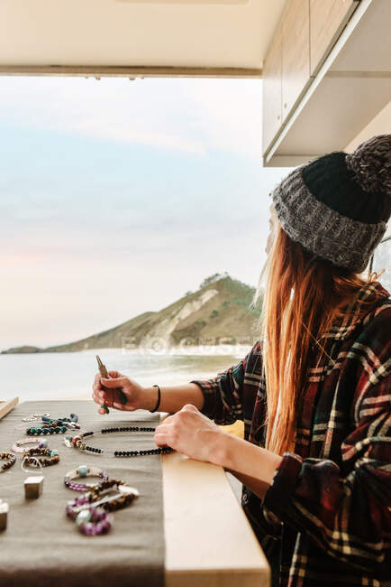 Side view of content female traveler creating handmade accessories while sitting at wooden table in parked truck at seaside — Stock Photo