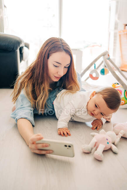 Игривая женщина делает селфи с очаровательным малышом на мобильном телефоне, проводя время дома — стоковое фото