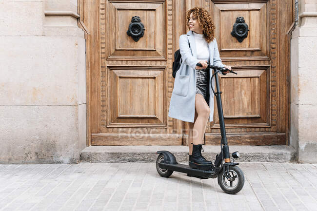 Full body of African American smiling young female in blue coat with scooter standing on city street near historic building and looking away - foto de stock