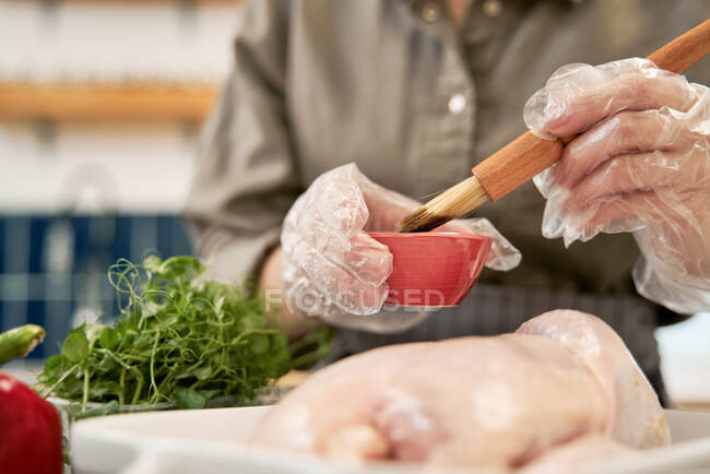 Анонимная женщина с кондитерской кисточкой, смазывающая сырую птицу соевым соусом во время приготовления пищи дома — стоковое фото