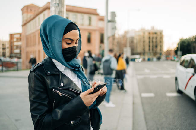 Vue latérale de la femme musulmane dans le hijab et la messagerie de masque de protection sur les médias sociaux sur le téléphone mobile tout en se tenant dans la rue de la ville — Photo de stock