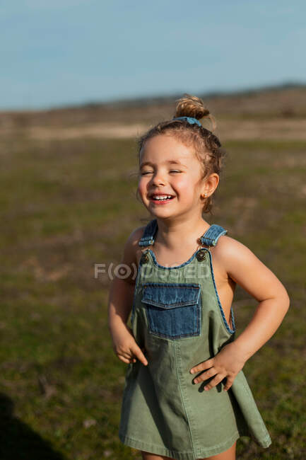 Зачарована чарівна маленька дівчинка в комбінезоні стоїть з руками на талії на лузі і дивиться вниз — стокове фото
