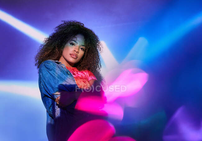 Attractive young African American female in casual wear with arms folded looking at camera while standing in dark studio in neon lights — Stock Photo