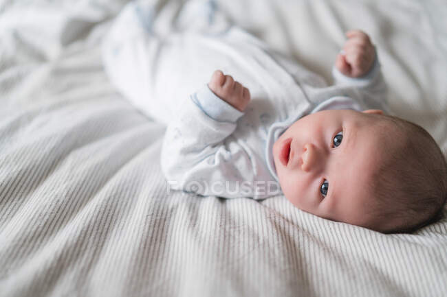 Очаровательный маленький ребенок в мягкой одежде лежит на измятой кровати, глядя в сторону дома — стоковое фото