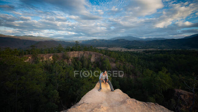 Мужской турист сидит на грубой скале, глядя в сторону от гор с деревьями в Таиланде — стоковое фото