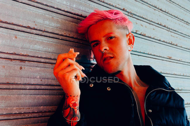 Junger homosexueller Mann mit Tätowierung und rosa Haaren in stylischer Oberbekleidung blickt vor verwitterter Wand in die Kamera — Stockfoto