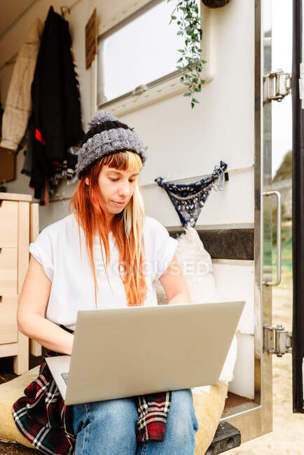 Delighted female traveler sitting outside on stairs of van while working remotely on laptop — Stock Photo
