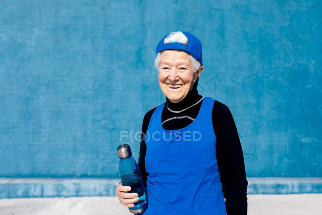 Desportista madura alegre em activewear e tampa de pé com garrafa de água na mão contra a parede azul no centro de treinamento ao ar livre ensolarado e olhando para longe — Fotografia de Stock
