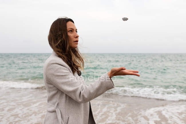 Vista laterale di donna calma in cappotto in piedi sulla spiaggia vicino al mare e gettando pietra nella giornata nuvolosa in autunno — Foto stock