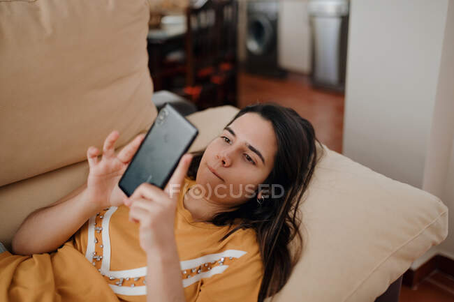Junge Frau schreibt SMS auf Handy, während sie sich auf Couch im Wohnzimmer hinlegt — Stockfoto