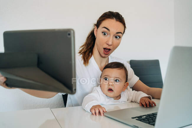 Etonné jeune mère et bébé mignon assis au bureau et parcourant netbook et tablette ensemble dans la salle de lumière — Photo de stock
